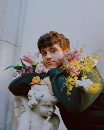Young adult male poses with a statue adorned with flowers. He has brown hair and a green top, near stone columns in natural light.