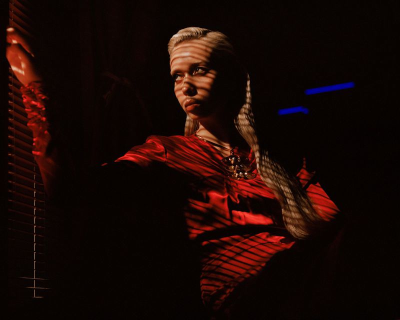 Portrait of a young woman in a red satin top looking out a window with blinds casting shadows across her face in a dark indoor space.
