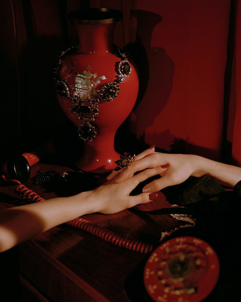 Still life composition featuring hands near a red vase adorned with a necklace and various red vintage objects on a wood surface.