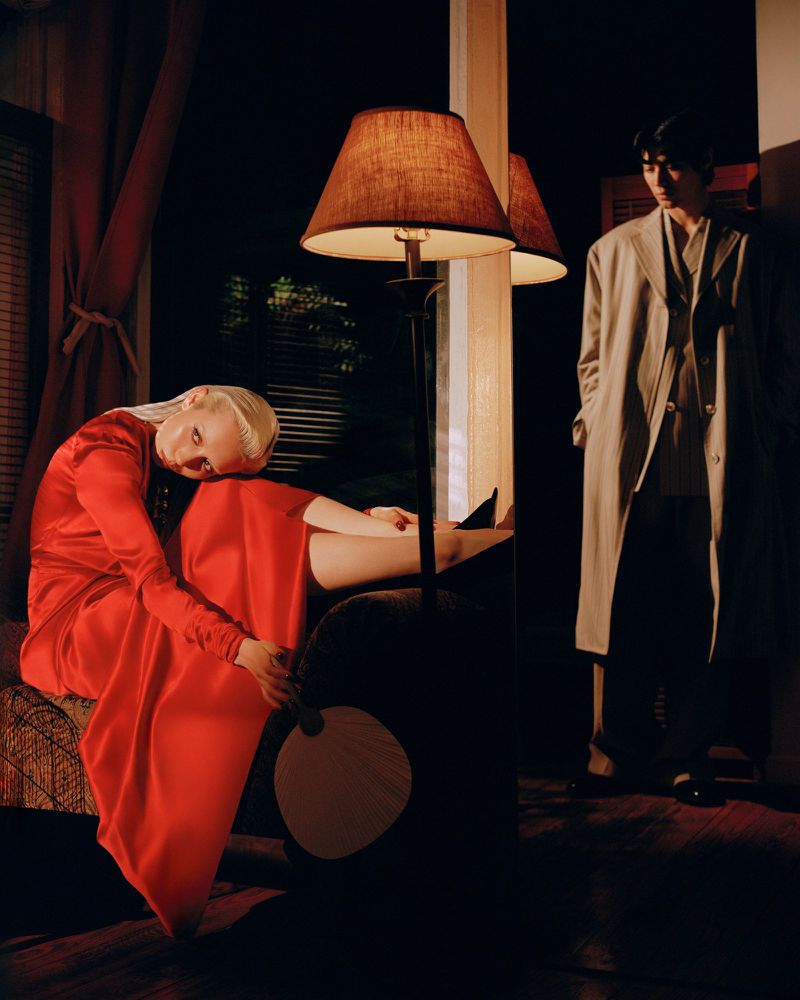 Woman in red dress sitting with legs resting on armchair next to floor lamp. Man in coat stands in the background.