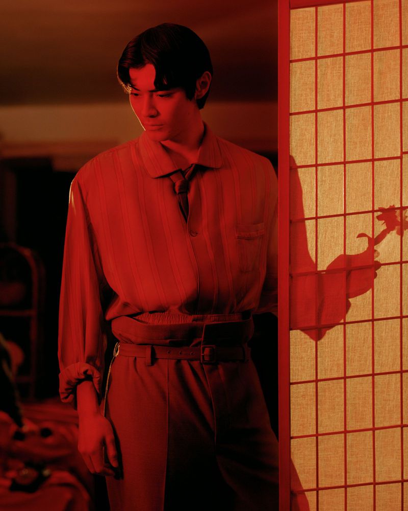 Stylish young adult stands beside a shoji screen in warm, red-toned light. Polo shirt and high-waisted pants create a vintage mood.