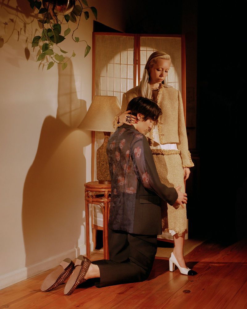 Blonde woman in a tan suit stands as a person kneels and embraces her leg in a dimly lit interior with plants and wooden floors.