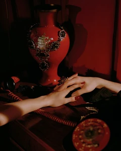 Close-up of hands touching on a wooden surface beside an ornate necklace on a red vase and a vintage telephone.