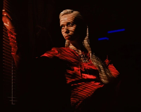 Model with textured blonde hair wearing a red satin blouse, illuminated by venetian blind shadows in dark low-key lighting.