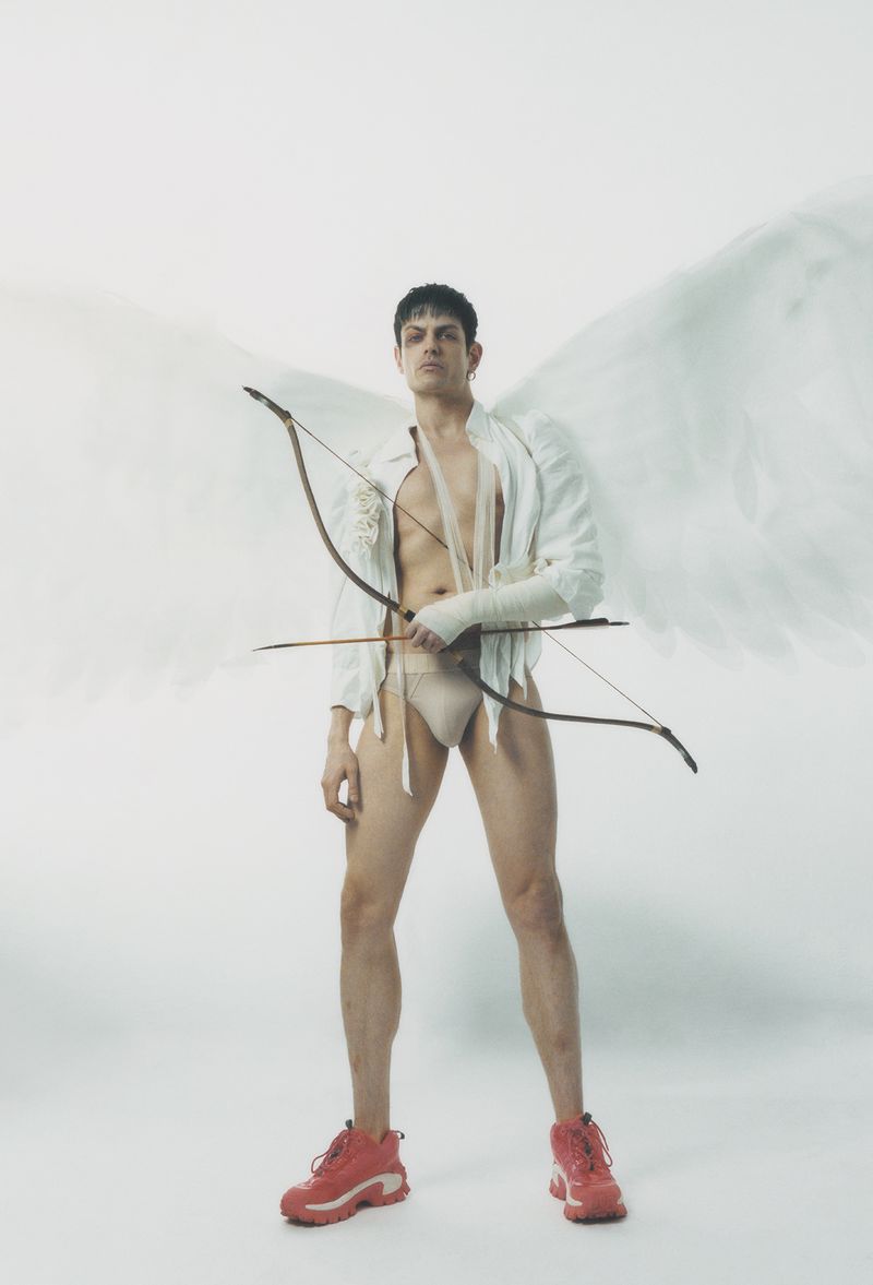 Man dressed as Cupid with wings, wearing trainers and a bow and arrow, posing against a white backdrop in a studio setting.