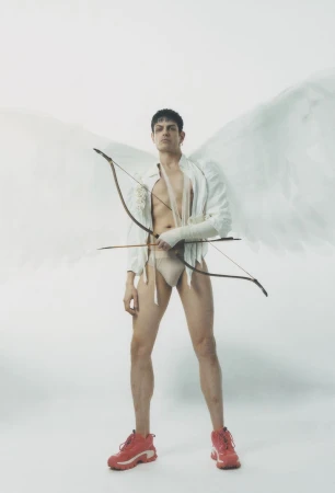 Man dressed as Cupid with wings, wearing trainers and a bow and arrow, posing against a white backdrop in a studio setting.