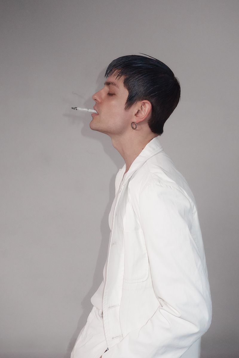 Side-profile shot of an adult male with short dark hair smoking a cigarette while wearing a white jacket in front of a grey wall.