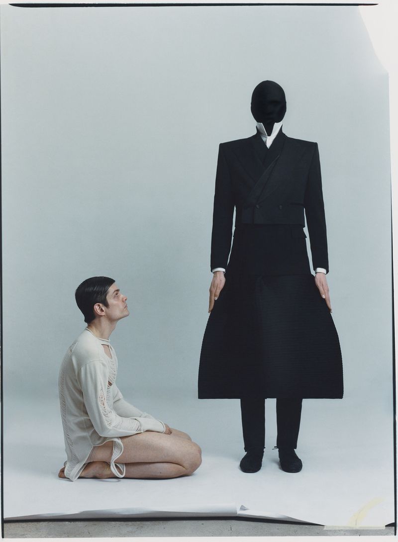 A man kneeling and looking up at a standing mannequin with a black mask and dark suit against a light backdrop in a studio shot.