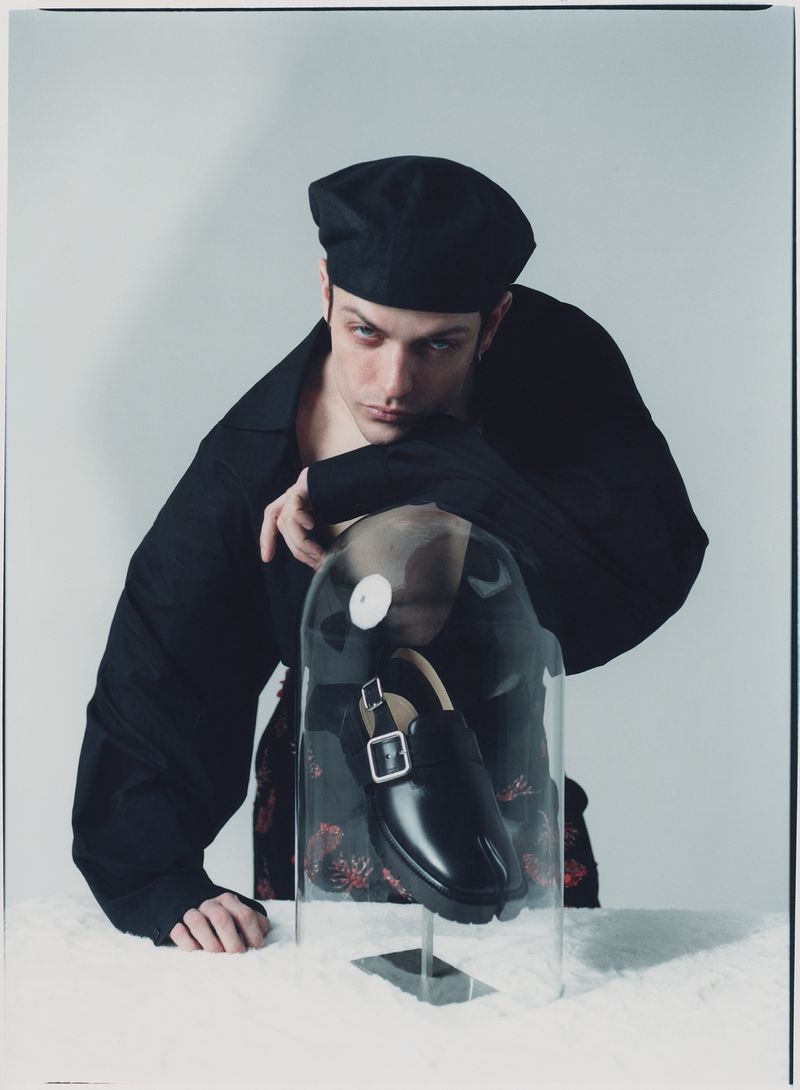 Fashion shot of a male model wearing a black beret and black jacket posed next to a shoe encased in clear plastic.