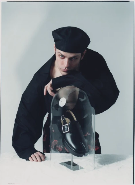 Fashion shot of a male model wearing a black beret and black jacket posed next to a shoe encased in clear plastic.