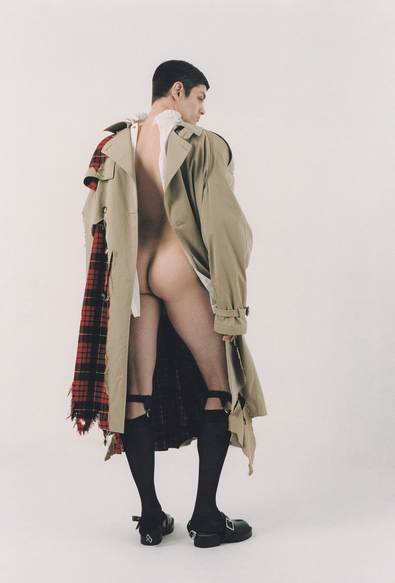 A nude male model poses in a photo studio wearing a beige trench coat, black stockings, and black Mary Jane shoes, with a white background.