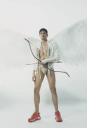 Model wearing white angel wings, unbuttoned shirt, briefs, and red sneakers holding a bow and arrow against a white background.