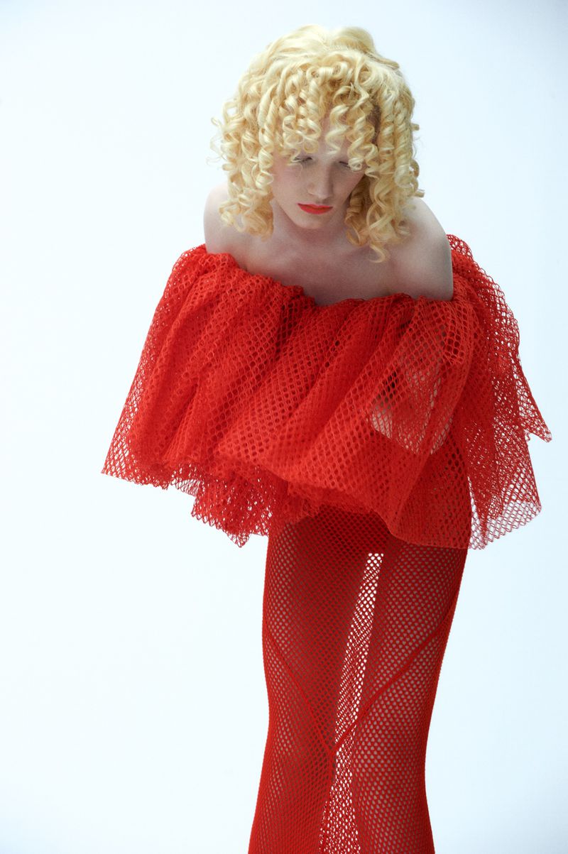Fashion shot of a blond woman in a red net dress, posing in a photo studio against a white background. Curly blond hair and bright red lipstick.
