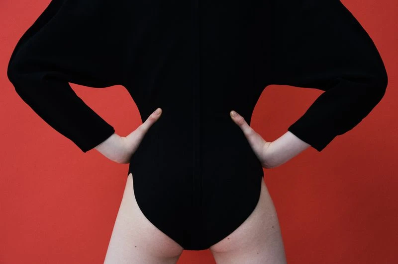 Woman in a black bodysuit posing with hands on hips against a red background in a studio, showing fashion style.