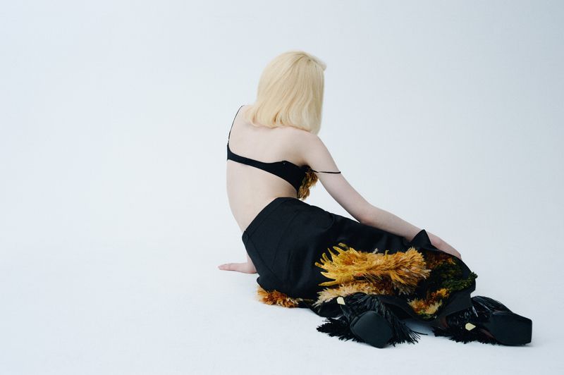 Woman with blonde hair wearing black top and black pants with feathers sits on white floor in a fashion studio setting.