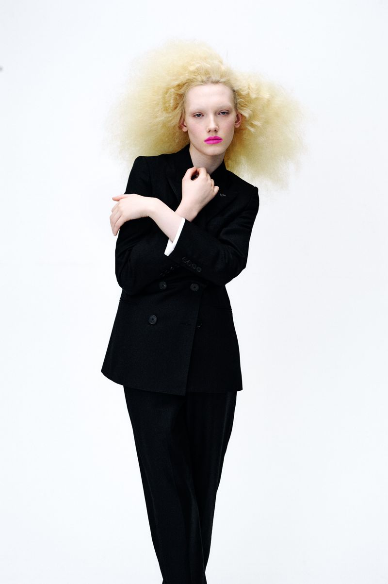 Woman with blonde afro poses in a black double-breasted suit against a white backdrop, showcasing fashion and modern style.