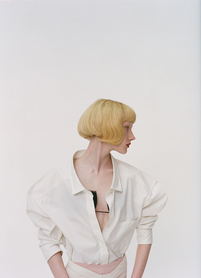A woman with a blonde bob hairstyle poses in a white collared shirt with a black bra strap showing in a light studio setting.