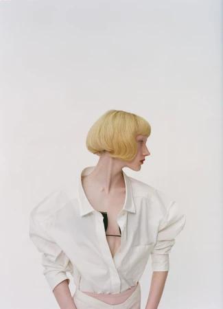 A woman with a blonde bob hairstyle poses in a white collared shirt with a black bra strap showing in a light studio setting.