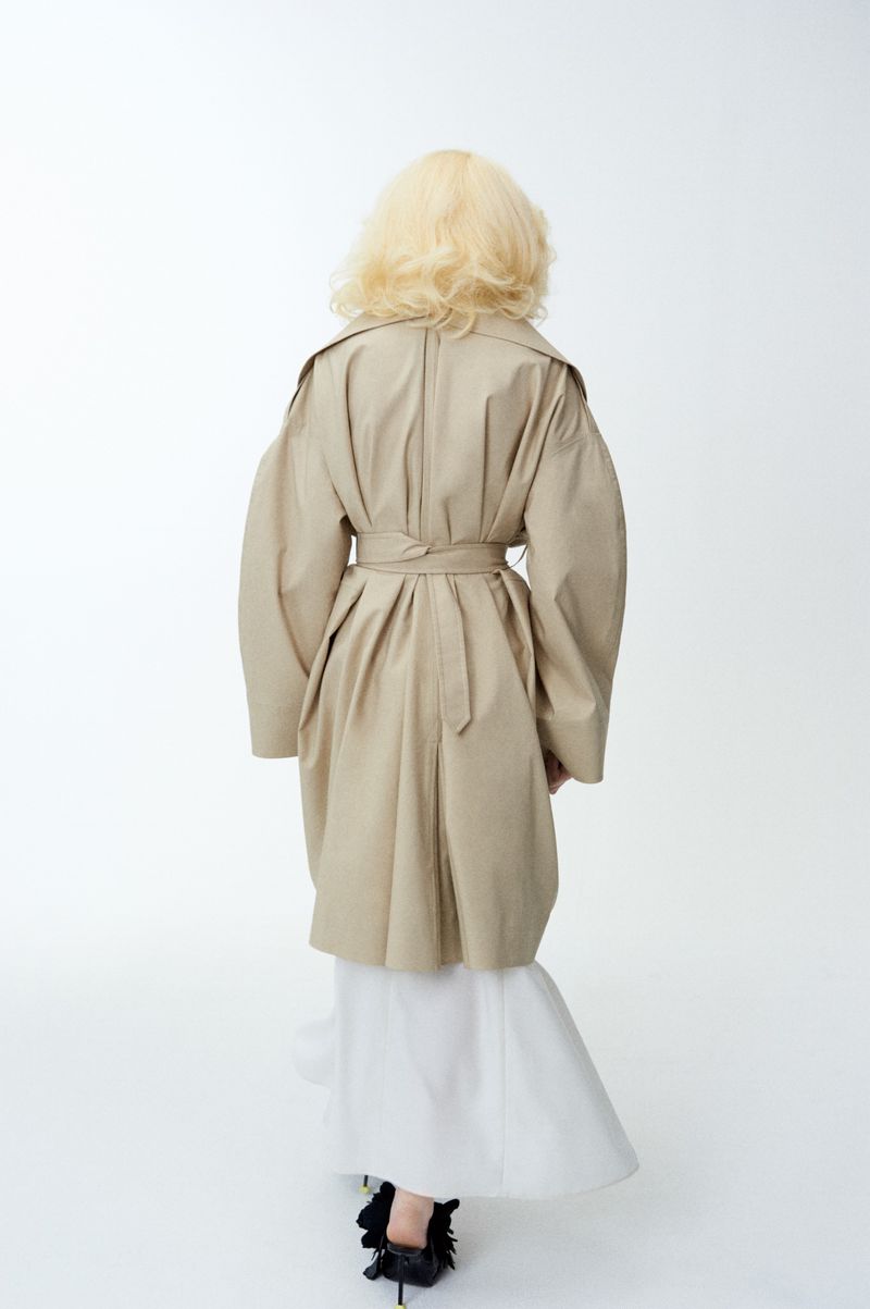 Back view of a blond woman in a tan trench coat and white dress, walking in a brightly lit photo studio with a soft, clean aesthetic.