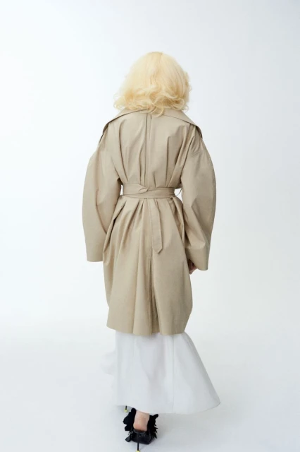 Back view of a blond woman in a tan trench coat and white dress, walking in a brightly lit photo studio with a soft, clean aesthetic.