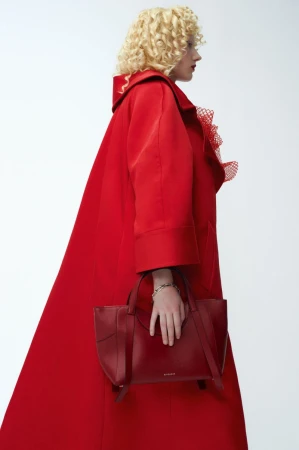 Model with curly blonde hair wearing an oversized red coat and holding a burgundy leather handbag against a white seamless background.