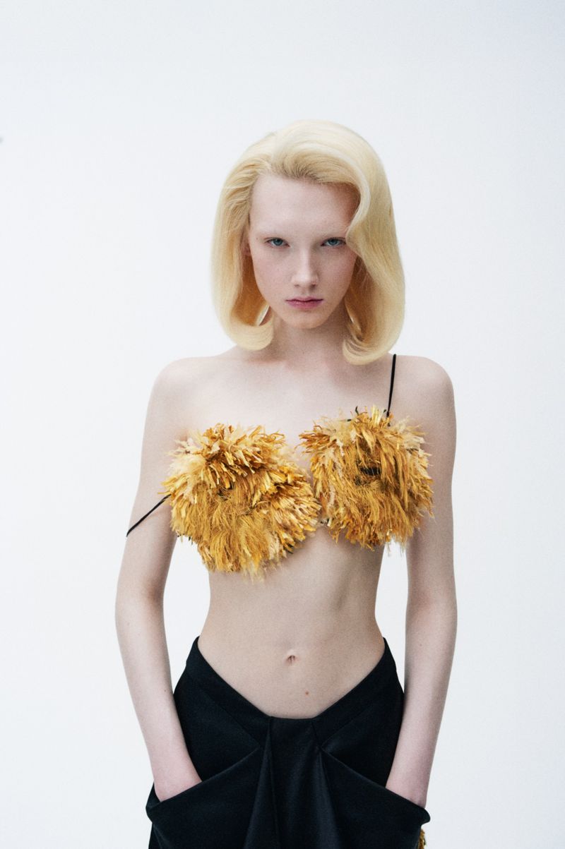 A woman with platinum blonde hair poses in a feather bra and black skirt against a bright white background in a studio shot.
