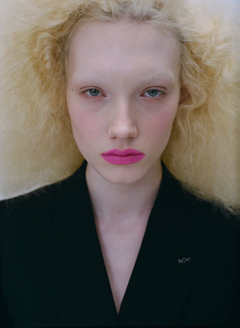 Close-up portrait of a fair-skinned adult female with bright pink lipstick and voluminous blonde hair wearing a black blazer against a white background.