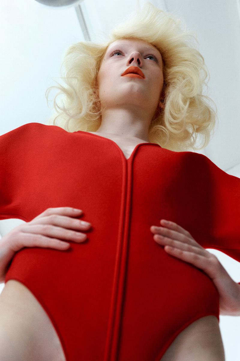 Blonde woman in a red bodysuit gazes upward in a fashion-forward, eye-level portrait against a bright white backdrop, creating an avant-garde look.