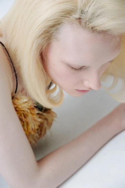 Close-up shot of a fair-skinned adult woman with blonde hair looking down; she is wearing a dress with feathered embellishments.