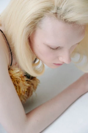 Close-up shot of a fair-skinned adult woman with blonde hair looking down; she is wearing a dress with feathered embellishments.