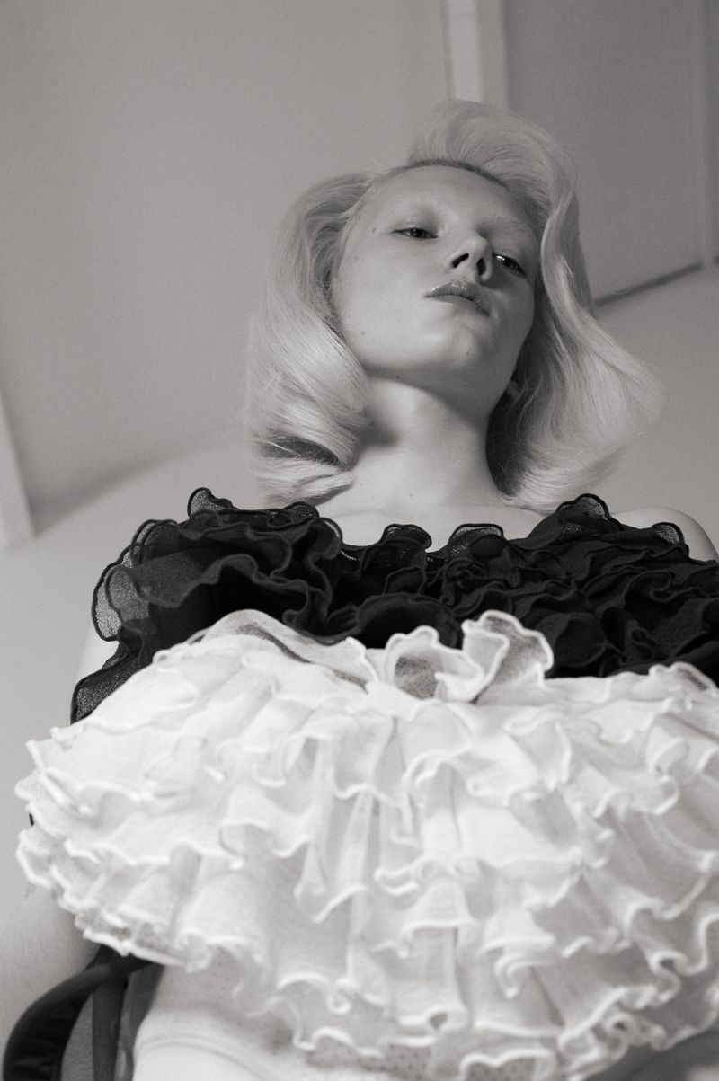 Monochrome portrait of a fair-skinned blonde woman in a ruffled collar, captured from a low angle in an indoor setting with soft lighting.
