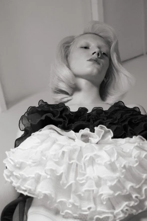 Monochrome portrait of a fair-skinned blonde woman in a ruffled collar, captured from a low angle in an indoor setting with soft lighting.