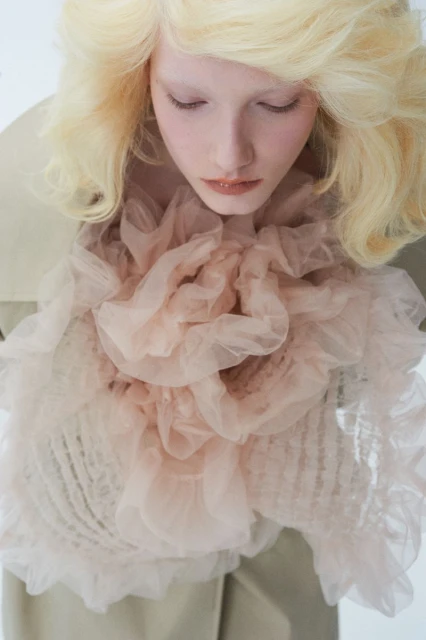 High-angle portrait of a fair-skinned woman with voluminous blonde hair, wearing a beige top and a large, ruffled, light-pink tulle collar.