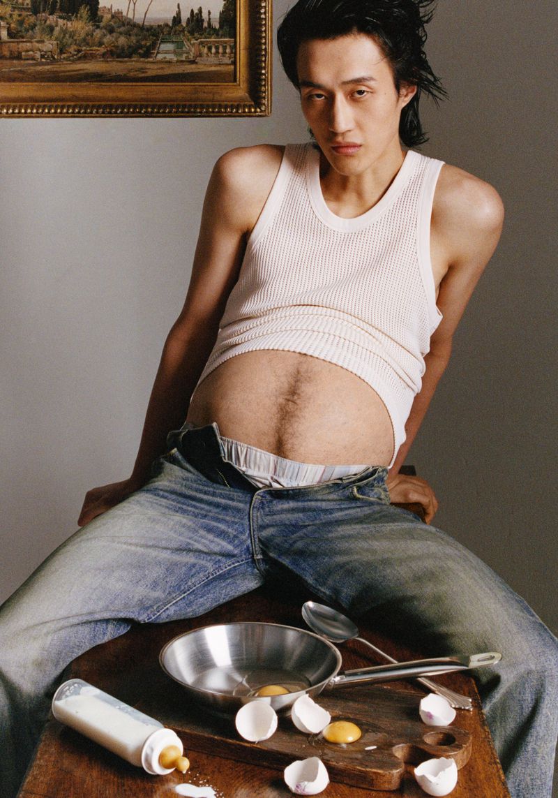 Adult Asian man poses with a frying pan of egg yolks, eggshells, and baby bottle on a wooden table in front of a light grey wall.