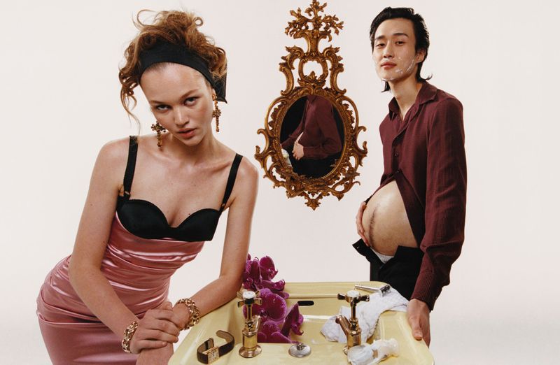 Fashion shot of woman in pink dress and man with shaving cream next to sink and mirror, evoking retro style and unique gender roles.