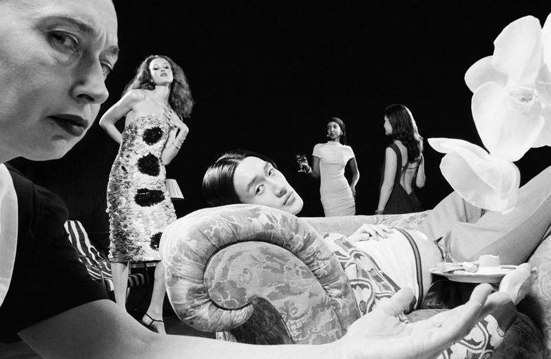 Black and white fashion shot with women in dresses, and a man reclining on a sofa, set against a dark background and orchids.