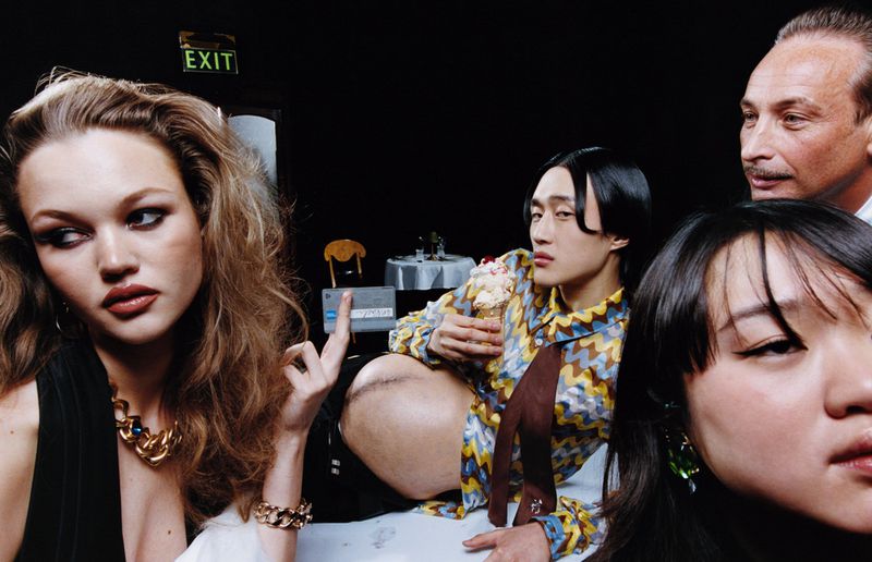 Group of people indoors with an Asian male eating ice cream, a blonde woman in a black top, and an exit sign visible in the background.