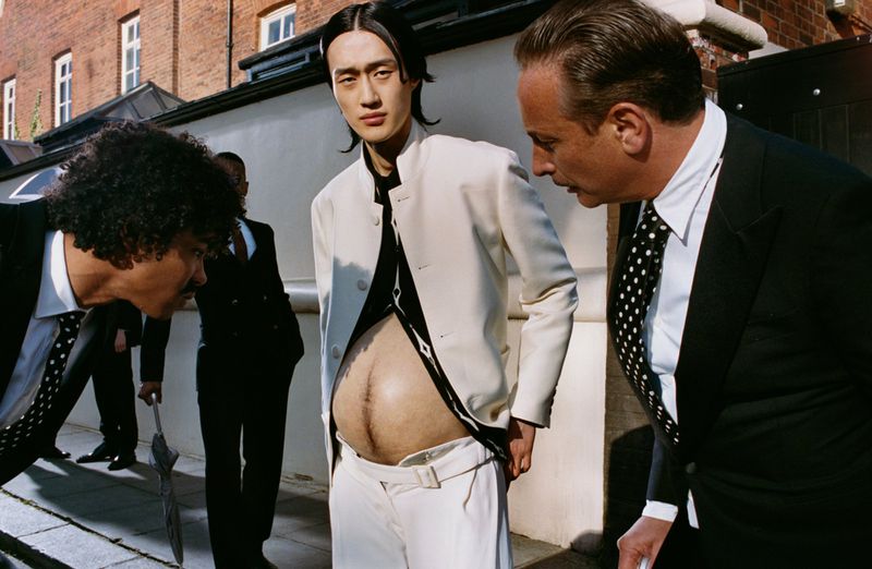Men in suits surround a person with an exposed prosthetic pregnant belly, photographed outdoors with a building in the background.