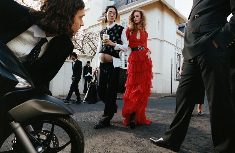 Fashion shoot featuring a group of people in formal wear standing on a city street with a motorcycle and architecture in the background.