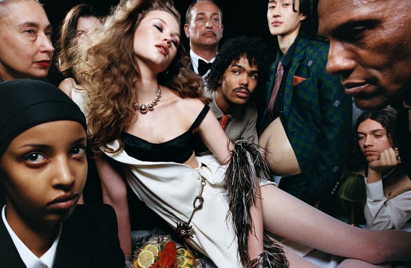 Fashion editorial shot of a group of diverse models in formal wear, surrounded by food and drinks in a dark indoor setting.