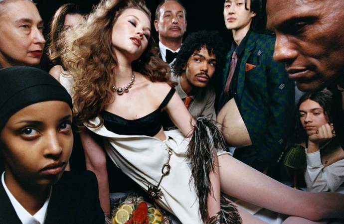 Fashion editorial shot of a group of diverse models in formal wear, surrounded by food and drinks in a dark indoor setting.