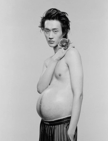 Monochrome studio portrait of a shirtless pregnant Asian man in striped pajama bottoms holding an orchid to his shoulder.