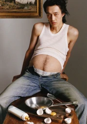 Man in white tank top and blue jeans seated behind a table with cracked eggs and a pan, photographed by Camille Summers-Valli for Dazed Magazine.