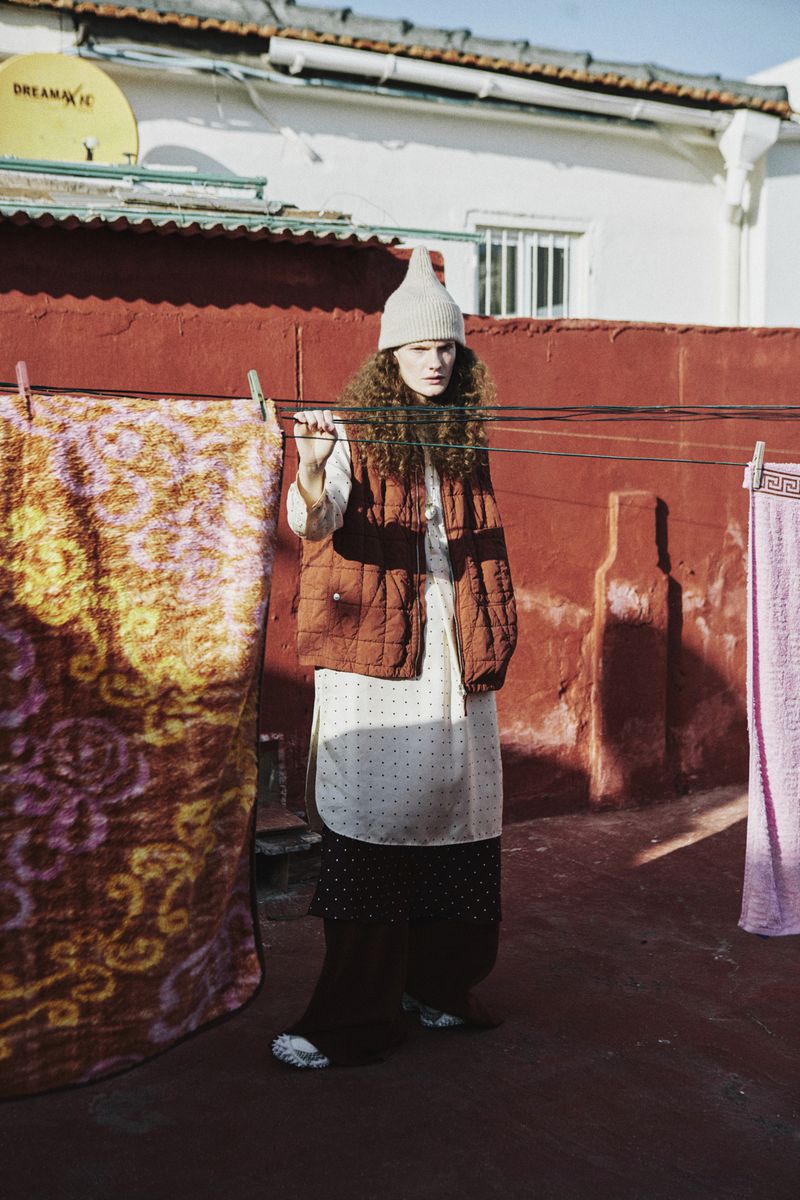 An adult female in a knit cap and vest stands outside in a white, patterned dress, brown pants, and silver shoes on a sunny day.