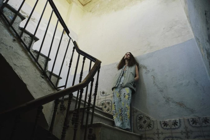 Adult woman in a long dress stands on a staircase indoors with handrails to the left side, looking up in a fashion style photograph.
