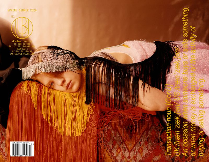 Magazine cover featuring a model with fringed headwear and clothing, lying on an ornate patterned surface, shot in soft, diffused light.