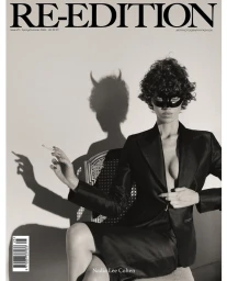 Nadia Lee Cohen wearing a black eye mask and tailored blazer seated on a chair with a horned shadow cast on the wall.