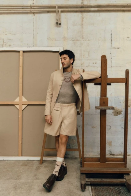 A man in a blazer and shorts poses next to an easel in a studio, with a neutral color palette and soft lighting.