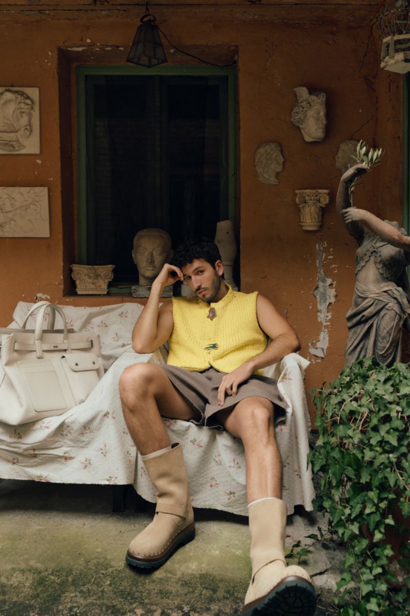 An adult male sits on a chair outside wearing a yellow sweater vest, brown shorts, and tan boots, posing next to a white bag and greenery.