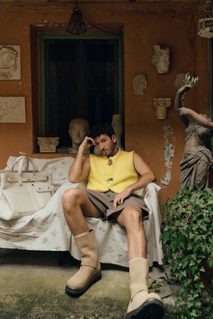 An adult male sits on a chair outside wearing a yellow sweater vest, brown shorts, and tan boots, posing next to a white bag and greenery.